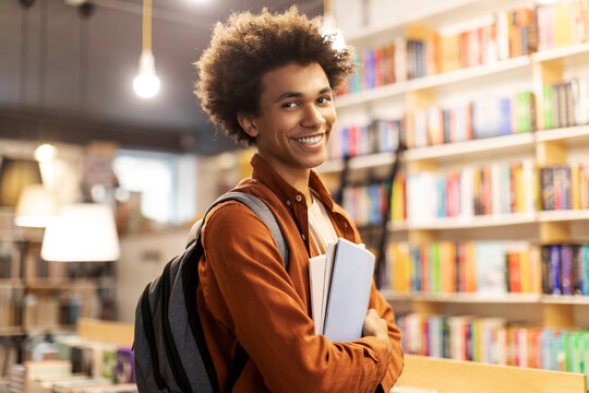 Portrait of happy African American guy wearing backpack and holding copybooks, smiling at camera at college library