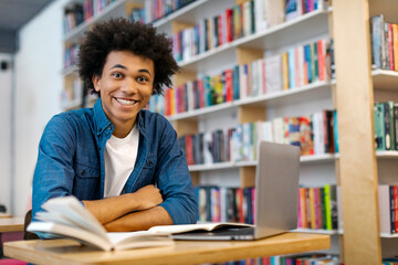 Portrait of excited African American male student sitting in university library at table with laptop computer and smiling at camera, free space