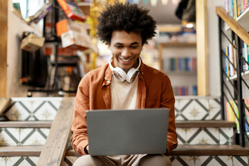 Happy black student guy using laptop while sitting on stairs in university library, having break...