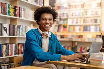 Happy African American male student sitting at desk in library smiling at camera, having online class or preparing for lecture