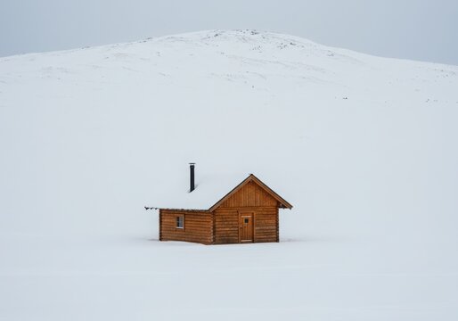 A small wooden cabin sits alone in a vast, snowcovered landscape under a cloudy sky