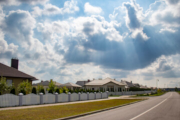 Blurred view of a quiet suburban neighborhood with private houses, white fences, and a curved street under a cloudy sky. Background for real estate, property, and urban development themes.