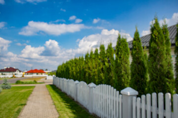 Blurred view of a quiet suburban neighborhood with private houses, white fences, and a curved street under a cloudy sky. Background for real estate, property, and urban development themes.