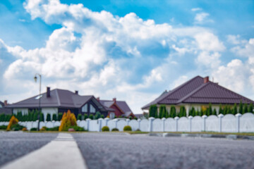 Blurred view of a quiet suburban neighborhood with private houses, white fences, and a curved street under a cloudy sky. Background for real estate, property, and urban development themes.