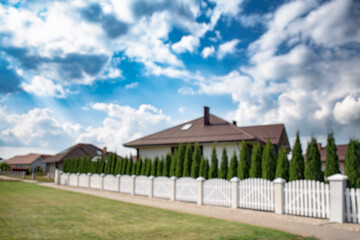 Blurred view of a quiet suburban neighborhood with private houses, white fences, and a curved street under a cloudy sky. Background for real estate, property, and urban development themes.