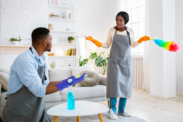 Fototapeta premium Quarrel over household chores. Angry black man and woman arguing while cleaning up their home, copy space. Millennial African American couple fighting over domestic duties