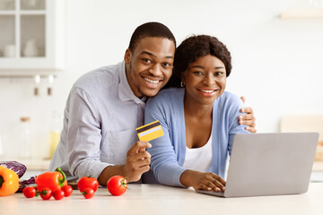 Happy black couple using laptop and credit card in kitchen, shopping online while cooking together....