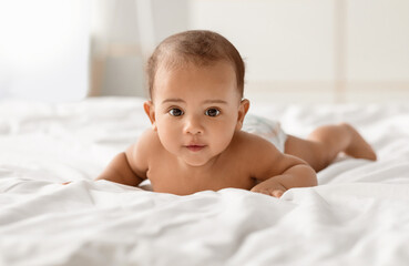 Children Beauty. Portrait of cute small African American baby crawling on bed on the white blanket...