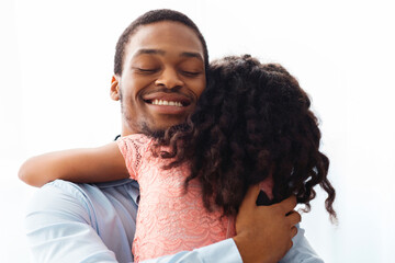 Ecstatic african american man hugging his little curly daughter, standing next to window at home,...