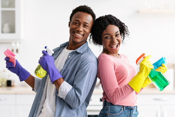 Joyful black couple with supplies for cleaning house posing in kitchen, funny african american man...