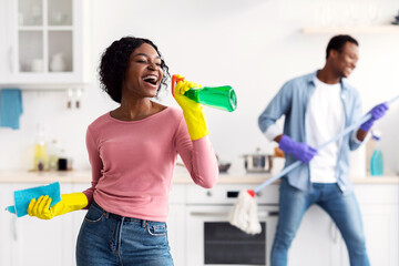 Joyful black lady having fun with her boyfriend while cleaning kitchen at home, african american...
