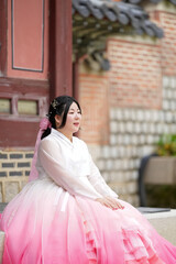 Naklejka premium In June in Seoul, a Korean woman in her twenties sits beside a historic building at Gyeongbokgung Palace, wearing a cute pink hanbok and admiring ornate details under a clear summer sky. Seoul, June.