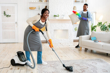 Happy black woman vacuuming floor while her boyfriend dusting cushion at their apartment, full...