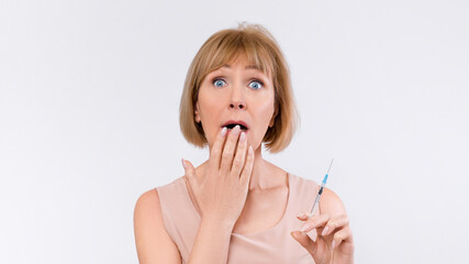 Shocked senior lady holding injection medicine to prevent aging on light studio background. Woman...
