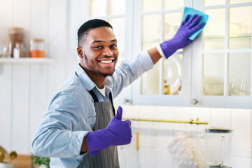 Portrait of young black guy in rubber gloves tidying kitchen cabinet and showing thumb up gesture...