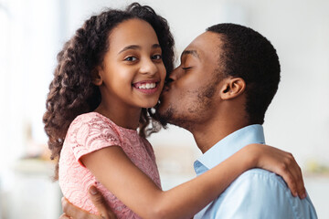 Handsome black father kissing his cute little daughter, closeup portrait, home interior. Adorable...