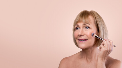 Best makeup for mature skin. Beautiful senior lady putting on natural cosmetics with brush over her...