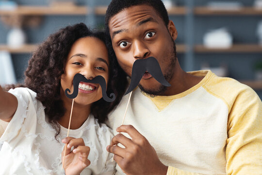 Funny african american father and daughter taking selfies together while staying home, using selfie accessories mustaches on sticks, looking at camera and smiling, closeup portrait