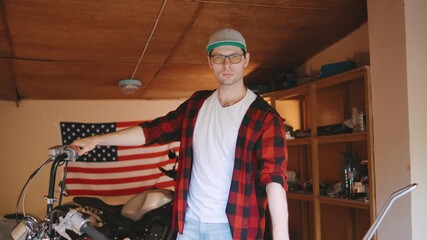 Portrait of biker man in a checkered shirt working on a motorcycle in a garage decorated with an American flag in the backdrop during daylight - Powered by Adobe