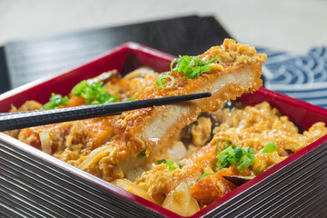 A lacquered Japanese bento box features a crisp pork cutlet glazed in savory sauce and garnished with spring onions, freshly picked up by chopsticks to highlight the dish in a menu photo.