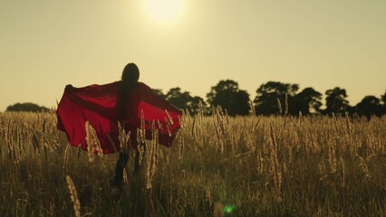 A girl runs across the field in a red superhero cloak waving in the wind at sunset, childhood dream...