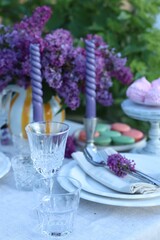 Beautiful lilac flowers, sweets, plates, cutlery, glasses and candles on table in garden, closeup