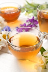 Aromatic herbal tea in glass cups and different flowers on white wooden table, closeup