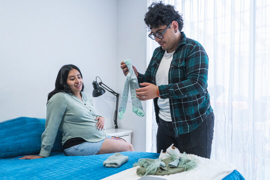 Happy pregnant woman watching her husband preparing baby clothes in bedroom