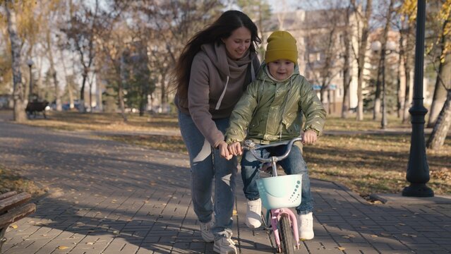 mother teaches little child to ride bike in the city, kid rides on sidewalk, happy family, baby plays and laughs with mom, learn ride, make childhood dream come true girls, daughter and mom are funny.