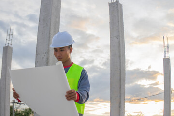 Confident young male engineer wearing hard hat, checking checklist and blueprints at construction...