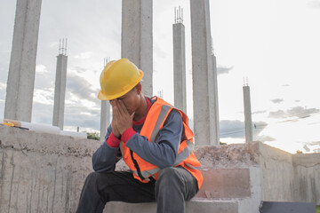 Male worker wearing safety suit and helmet sits on a ladder, showing fatigue and stress from hard...