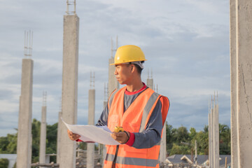 Male engineer in hard hat and safety vest examines blueprint indoors at construction site; planning...
