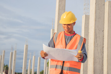 Male engineer in hard hat and safety vest examines blueprint indoors at construction site; planning...