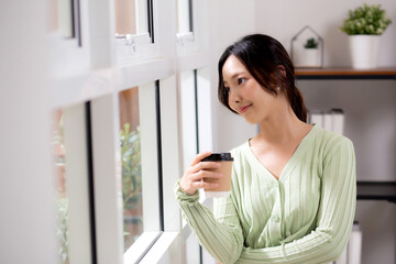 Portrait young asian woman relax and enjoyment with drinking coffee by the window in living room at home, woman smiling with happiness and comfortable drinking beverage at home, lifestyle and leisure.