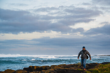 Obraz premium Scenic view of a man walking on the rocks at sea shore with tides and cloudy sky in the background, Griffiths Island, Port Fairy, Great Ocean Road, Victoria, Australia