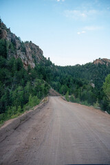 Remote dirt road winds through a rocky mountain pass surrounded by dense green forest under a clear blue sky.