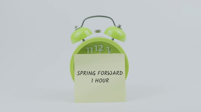 Spring Forward 1 Hour word written on a sticky note placed in front of a green ringing clock on white background, symbolizing daylight saving time, clock adjustment, and schedule change concept