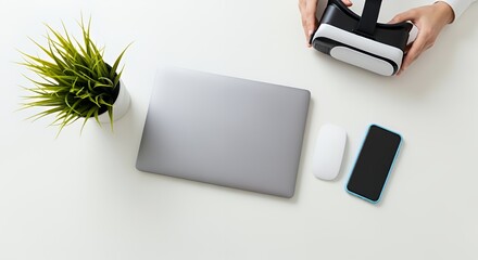 A person holds a virtual reality headset near a laptop, smartphone, and mouse on a white desk, showcasing modern technology and workspace essentials.