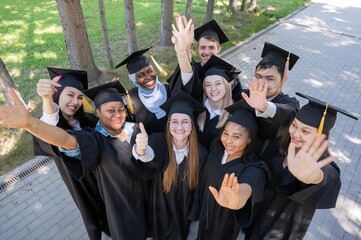 Students of different nationalities in graduation gowns give thumbs up. 