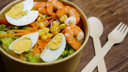 Healthy and colorful salad featuring shrimp, boiled eggs, lettuce, carrots, corn, and red cabbage in a disposable bowl with wooden cutlery, highlighting balanced eating and eco friendliness