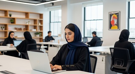 A young woman in a hijab works diligently on her laptop in a modern, shared workspace, surrounded by other professionals.