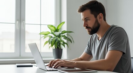 A focused young man works diligently on his laptop computer at home, enjoying the natural light from a nearby window.