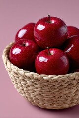 A rustic woven basket overflowing with fresh, glossy red apples on a solid pink background.