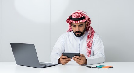A businessman in traditional Middle Eastern attire uses a tablet computer while working at his desk with a laptop.