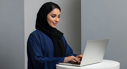 A young woman in a hijab works on her laptop, focused and smiling, in a modern, minimalist workspace.