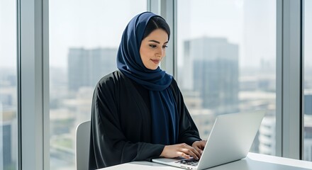 A young woman in traditional Middle Eastern attire works productively on her laptop in a modern, bright space overlooking a cityscape.