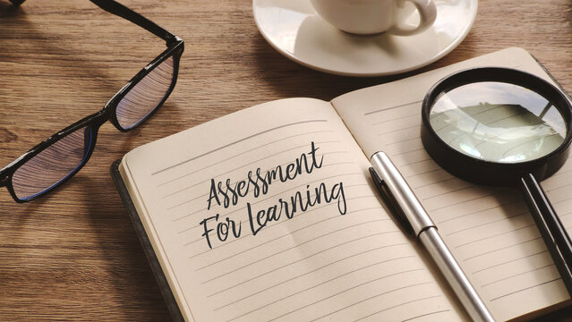 Top-down view of a wooden desk with an open notebook showing "Assessment For Learning," a pen, magnifying glass, glasses, and a cup of coffee.