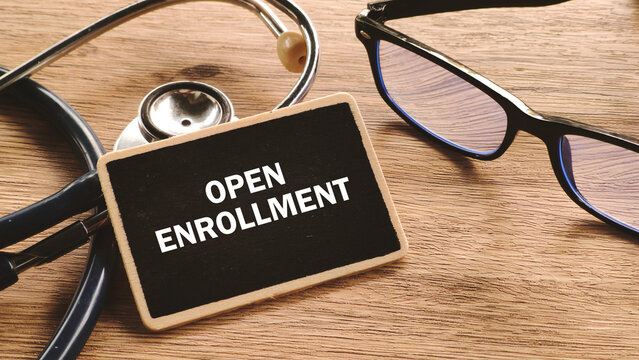 Top-down view of a wooden desk with a stethoscope, glasses, and a small chalkboard displaying "OPEN ENROLLMENT."