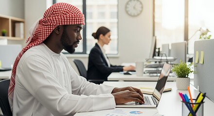 A man in traditional Middle Eastern attire works on his laptop in a modern, shared space, while a woman works at a nearby desk.