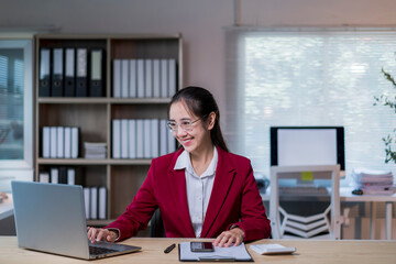 woman in a red jacket is sitting at a desk with a laptop and a cell phone